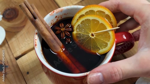 Warm Spiced Alcoholic Beverage Known as Mulled Wine Served in Red Colored Mug with Cinnamon Sticks, Orange Slices, and Cloves Resting on Wooden Surface