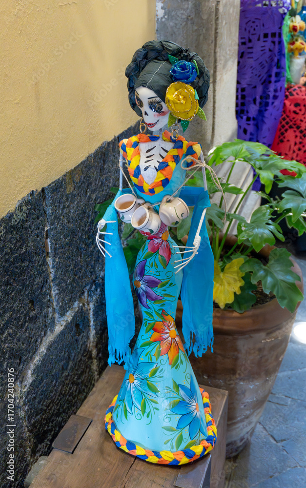 Fototapeta premium A skeleton dressed in a blue dress with flowers and a flower crown. The skeleton is holding a cup in one hand. Day of the Dead celebration in Mexico, papel picado crafts, offerings, La Catrina