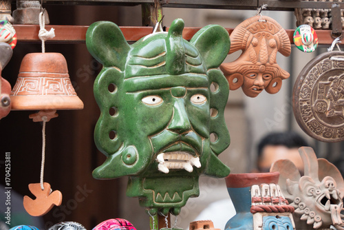 A green mask with a face on it is hanging on a wall. There are other masks and items hanging on the wall as well. Day of the Dead celebration in Mexico, papel picado crafts, offerings, La Catrina