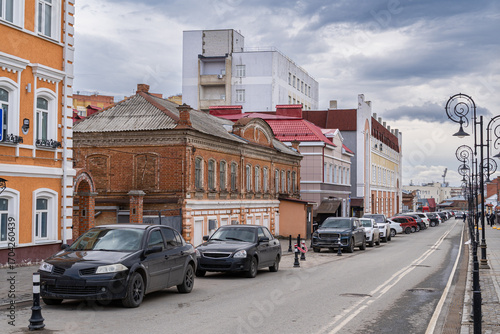 City street with parked cars and buildings of varying architectural styles. Brick and modern facades under a cloudy sky create a contrast between old and new.