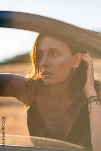 A girl in a black dress poses beautifully near an old car. The sun shines on the beautiful face of the girl who poses near an old car