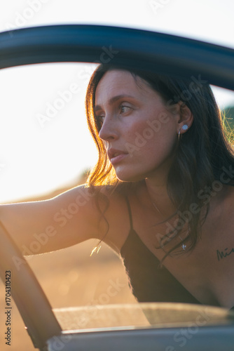 A girl in a black dress poses beautifully near an old car. The sun shines on the beautiful face of the girl who poses near an old car