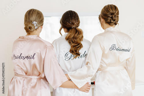 Bride in a satin robe with the inscription “Bride” on the back, preparing for her wedding day. Elegant white fabric with lace or embroidered details, captured in natural light.