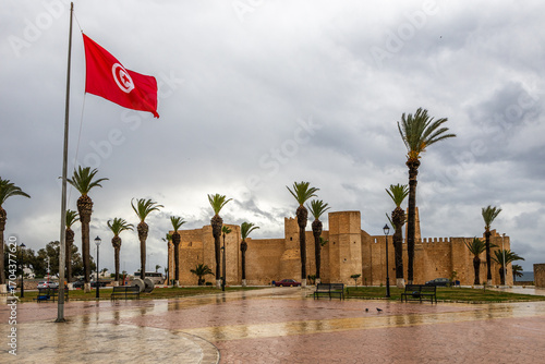 Photography Tunisian national flag with lots of palms and Ribat of Monastir under cloudy ski