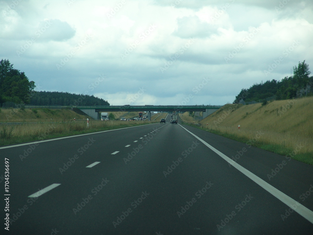 Fototapeta premium Multi-lane highway under cloudy skies with cars in motion, grassy median, and forested surroundings—modern road infrastructure in a natural setting.