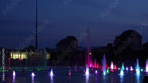 Spectacular colorful water fountain show with purple, pink and green LED lighting at night in city park with illuminated buildings silhouettes. Concept of urban entertainment and nighttime tourism.