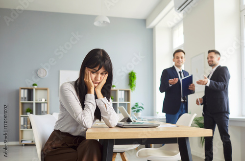 Foto Thoughtful female employee sits alone at a desk while two coworkers chat in the background, hinting at gossip and exclusion in an office