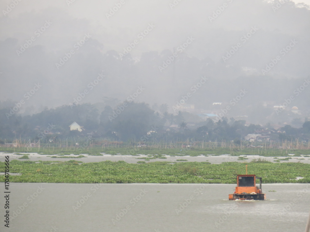 Fototapeta premium An orange machine boat is floating on the lake, designed to collect and clean water plants, with floating houses and bamboo structures in the background.