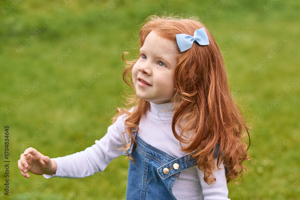 Obraz premium Young caucasian girl with red hair and blue bow smiling outdoors in green park setting.