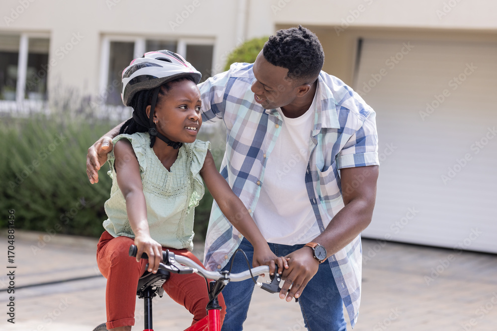 Fototapeta premium African American father teaching daughter to ride bicycle, both smiling outdoors