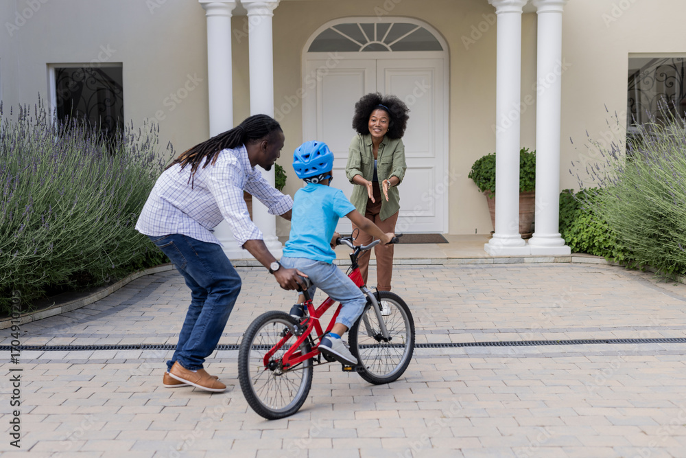 Naklejka premium African American parents teaching child to ride bicycle in front of house, smiling