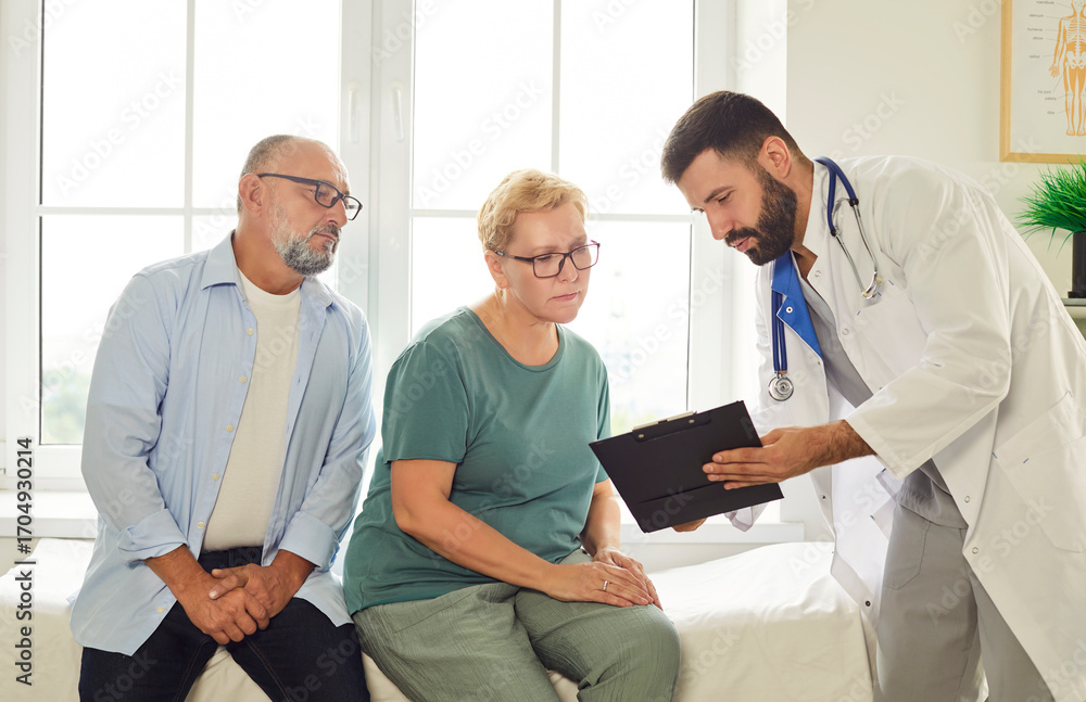 Obraz premium Family doctor reviews medical records with a senior couple in a clinic, explaining treatment options during a private consultation. Male physician talking with patients during examination. 