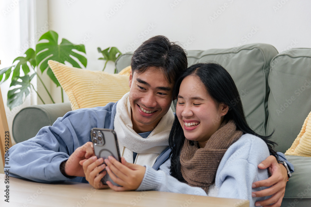 © sorapop - Winter day, happy couple sharing love at home, enjoying cozy inside warmth, wearing scarves, looking at phone, smiling, finding joy in digital content during cold winter season