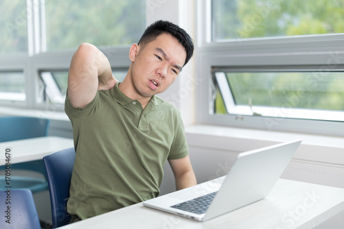 Young male student stretches and touches his neck in discomfort while using a laptop in a classroom setting, illustrating poor posture or tech related strain.