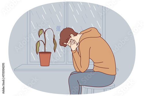 Depressed man sits alone and suffers from melancholy in room with window and wilted flower. Melancholy in older guy victim of autumn blues or everyday problems and troubles at work