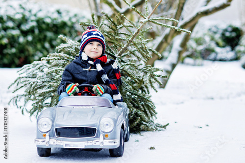 Funny little smiling kid boy driving toy car with Christmas tree.