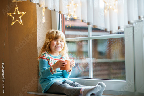 Little preschool girl holding cup with hot chocolate with marhsmallows. Happy child drinking sweet cocoa by window with Christmas lights in winter. Cozy family celebration of xmas.