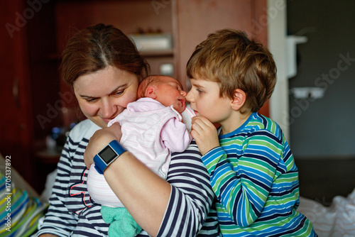 Happy little boy kissing his sleeping newborn sister in hospital bed. Mother holding baby girl on arm. Real life, real people moment of sibling love, bonding, and family tenderness