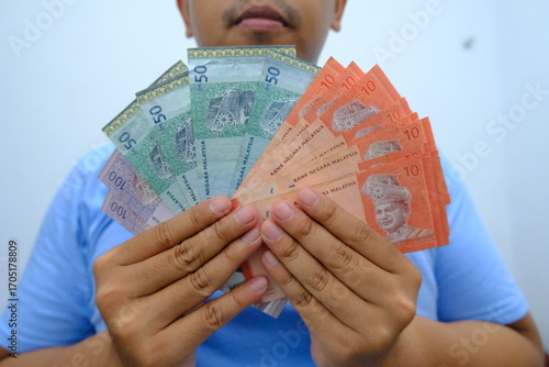 Close-up of a man in blue shirt holding Malaysian ringgit banknotes spread in a fan shape, symbolizing wealth and finance.  
