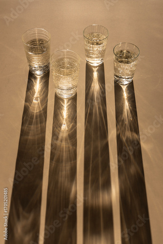 Four glasses of sparkling water casting long shadows on a beige background, light and shadows art