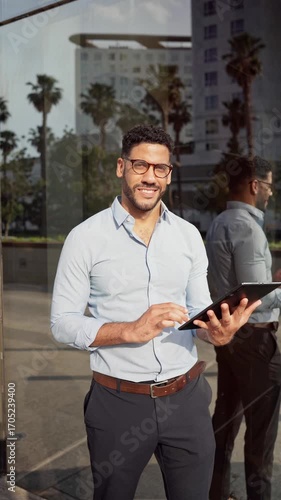 Vertical video of young Hispanic Businessman using tablet near modern building smiling at camera