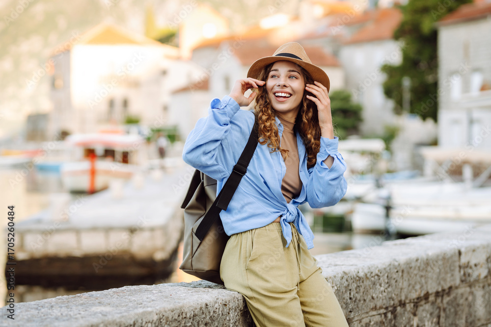 Fototapeta premium Portrait of a young woman wearing a hat and carrying a briefcase strolling along the sea in a picturesque town. The traveler enjoys the seascape. Concept of tourism and enjoyment.