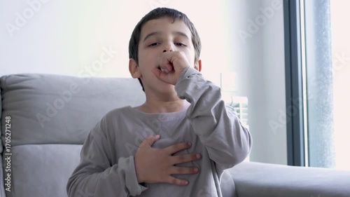 Child sitting on sofa at home and holding hand on chest. Male having asthma attack, difficulties with breathe, feeling severe pain or dyspnea