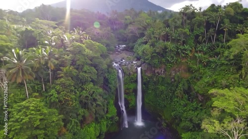 Twin waterfalls cascade into a basin, surrounded by a lush tropical forest and distant mountain under overcast skies