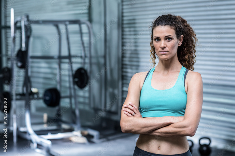 Fototapeta premium Female trainer standing arms crossed in gym by squat rack wearing workout attire, copy space