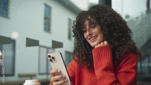 Caucasian woman with curly hair smiling while looking at smartphone in hand, sitting indoors near window. Female in red sweater drinking takeaway coffee and browsing phone at cafe table.
