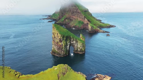 Aerial view of the majestic Drangarnir sea stack archway with vibrant green grass contrasting with the deep blue ocean waters, Drangarnir, Faroe Islands.