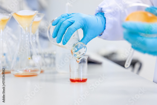 Close-up of hands pouring an orange chemical solution into a beaker, representing chemical analysis, food safety testing, and quality control procedures in a clean and professional lab environment.