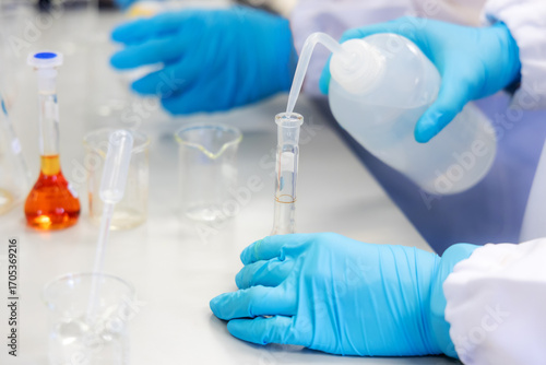 Wallpaper Mural A scientist carefully pours liquid from a laboratory wash bottle into a glass test tube during a chemical experiment. The modern laboratory with various glassware and scientific equipment visible. Torontodigital.ca