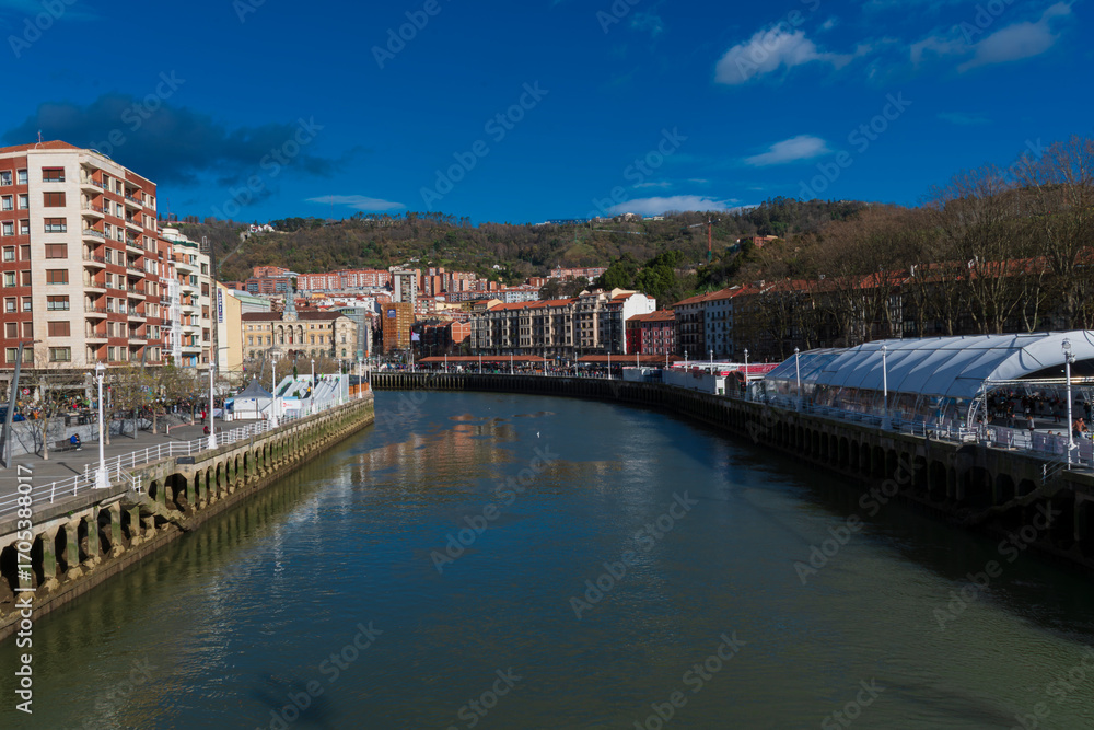 Obraz premium View of the Bilbao river with buildings and green hills in the background.