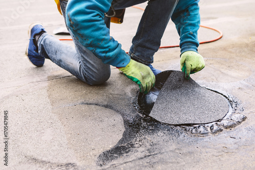 Roofer repairing a damaged section of a roof. Worker kneels on a wet, flat rooftop to apply a circular patch. Their gloved hands press the black material into place, sealing a roof