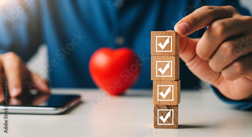 Person stacking wooden blocks with checkmarks near a phone and a red heart shaped object on a table