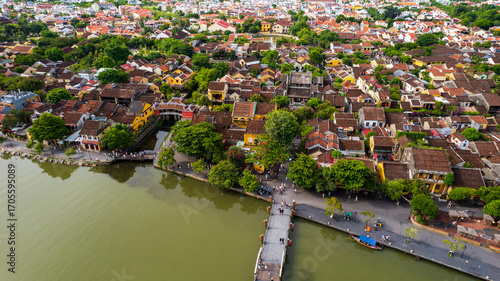 An aerial view of Hoi An's old town and river, showing the rows of historic houses and a long line of tourist boats, with a scenic backdrop of mountains and the modern city.