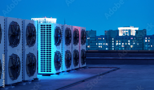 Фотография Rooftop industrial air conditioning units with large fans at night, cooling system for buildings, illuminated city skyline in background