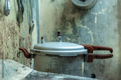 An Old alumunium pressure cooker with worn handles placed in a rustic kitchen, showing vintage cooking equipment against a weathered textured wall background