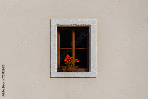 ein winzig kleines Sprossenfenster in der Mitte einer hellbeigen Fassade mit einem Blumentopf und Geranien auf der Fensterbank