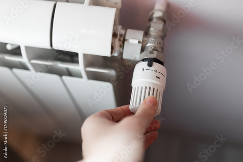 Closeup hand adjusting white plastic radiator thermostat knob set to zero with frost protection symbol, mounted on radiator with metal valve connection, household detail of energy saving temperature