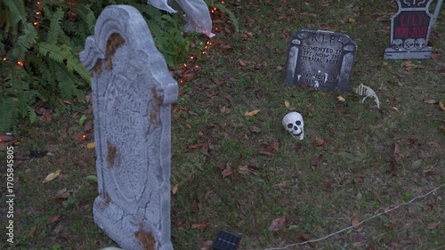 Outdoor Halloween scene with foam tombstones, a skull, bones, and orange lights on a grassy lawn. Spooky yard cemetery decoration with autumn leaves and ferns.