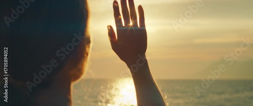 Person raising hand toward bright sunset over calm ocean horizon in the evening