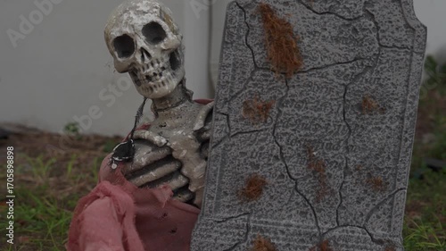 Close-up of a Halloween skeleton with a skull and bones leaning beside a cracked tombstone on a grassy lawn. Spooky outdoor cemetery decoration with tattered fabric, weathered stone, and autumn yard