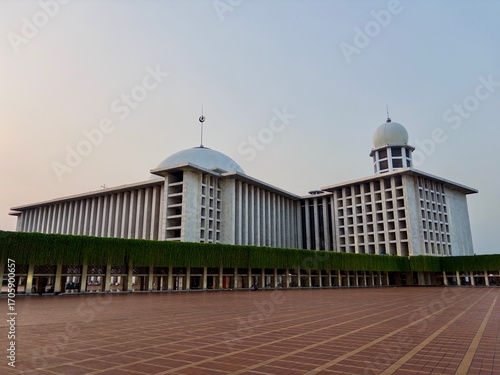 wide, symmetrical shot of the modern exterior of Istiqlal Mosque. The building's unique design and the green, hanging vines of its facade are beautifully highlighted under a clear sky.