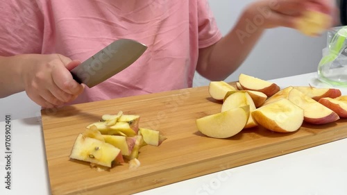A woman slices into a fresh red apple on a wooden cutting board, using a silver knife in a bright kitchen