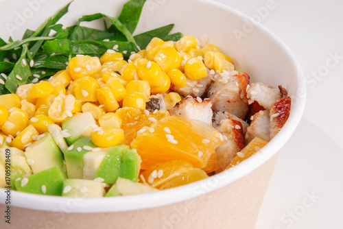 Bright poke bowl with juicy chicken pieces, fresh avocado, sweet corn, orange slices and greens, topped with sesame seeds, served in eco paper bowl, top view