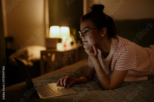 A young woman with glasses is lying on her stomach in bed, focused on her laptop in a dimly lit room. Soft light from a nearby lamp creates a cozy atmosphere.