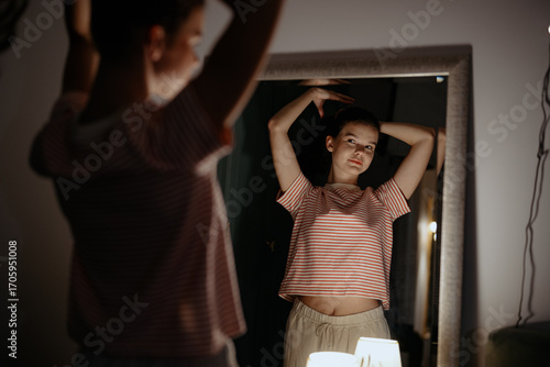 Fototapeta A young woman stands in front of a mirror, arms raised, as she admires her reflection in a softly lit room