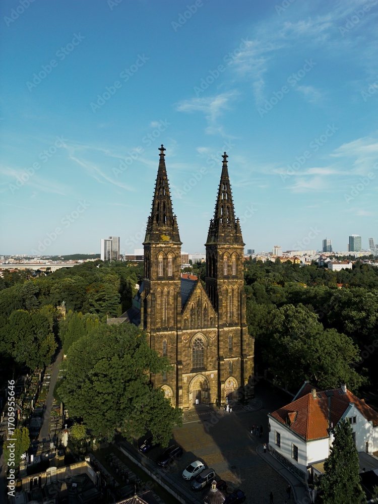 Naklejka premium Twin Gothic spires of the Vyšehrad basilica rising above the trees with city skyline in background.